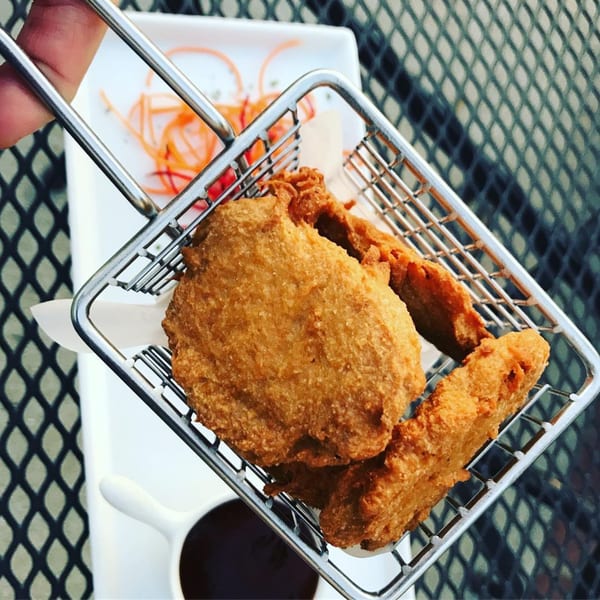 Crispy vegan fried chicken pieces in a metal basket, with a dipping sauce and pickled vegetables.