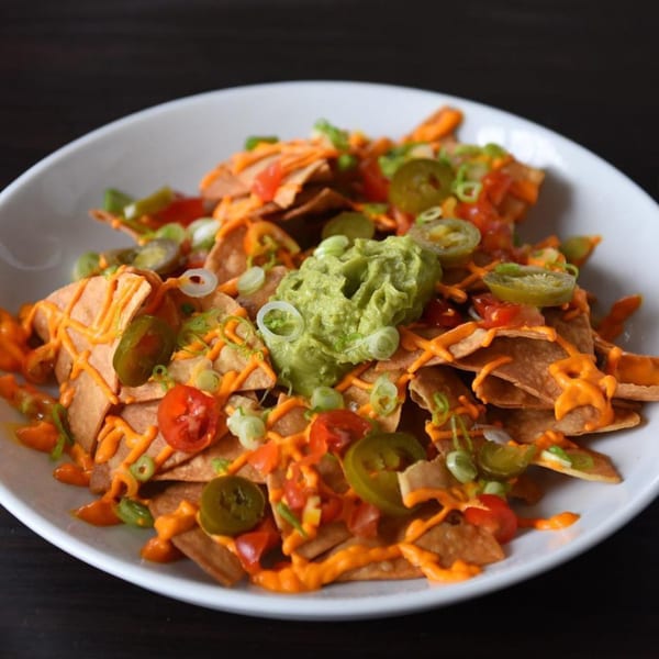 Vegan nachos with cheese sauce, guacamole, jalapeños, tomatoes, and green onions in a white bowl.