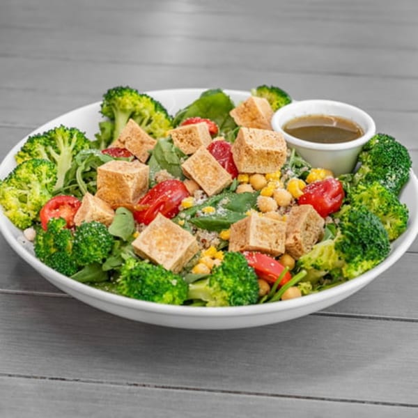 A vibrant vegan salad with broccoli, tofu, tomatoes, chickpeas, and a side of dressing.